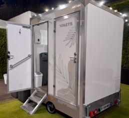 Caravane sanitaire mobile ouverte, avec accès par marche, lave-mains intégré et porte décorée de motifs floraux.