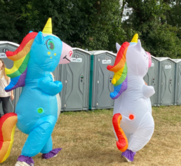 Deux carnavaleux déguisés en licornes gonflables marchent devant des cabines de toilettes mobiles WC Loc lors d’un carnaval.
