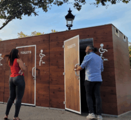 Deux personnes utilisent des toilettes publiques en bois, accessibles aux personnes à mobilité réduite, installées en extérieur dans un espace urbain arboré.