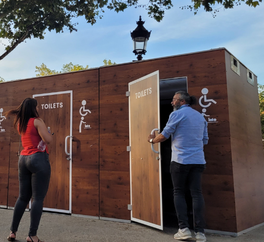 Deux personnes utilisent des toilettes publiques en bois, accessibles aux personnes à mobilité réduite, installées en extérieur dans un espace urbain arboré.