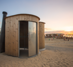 Toilette sèche en bois ronde installée sur une plage au coucher du soleil, avec bar de plage animé en arrière-plan