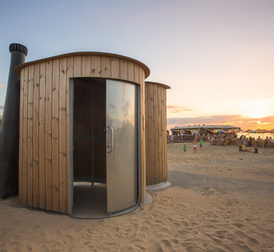 Toilette sèche en bois ronde installée sur une plage au coucher du soleil, avec bar de plage animé en arrière-plan